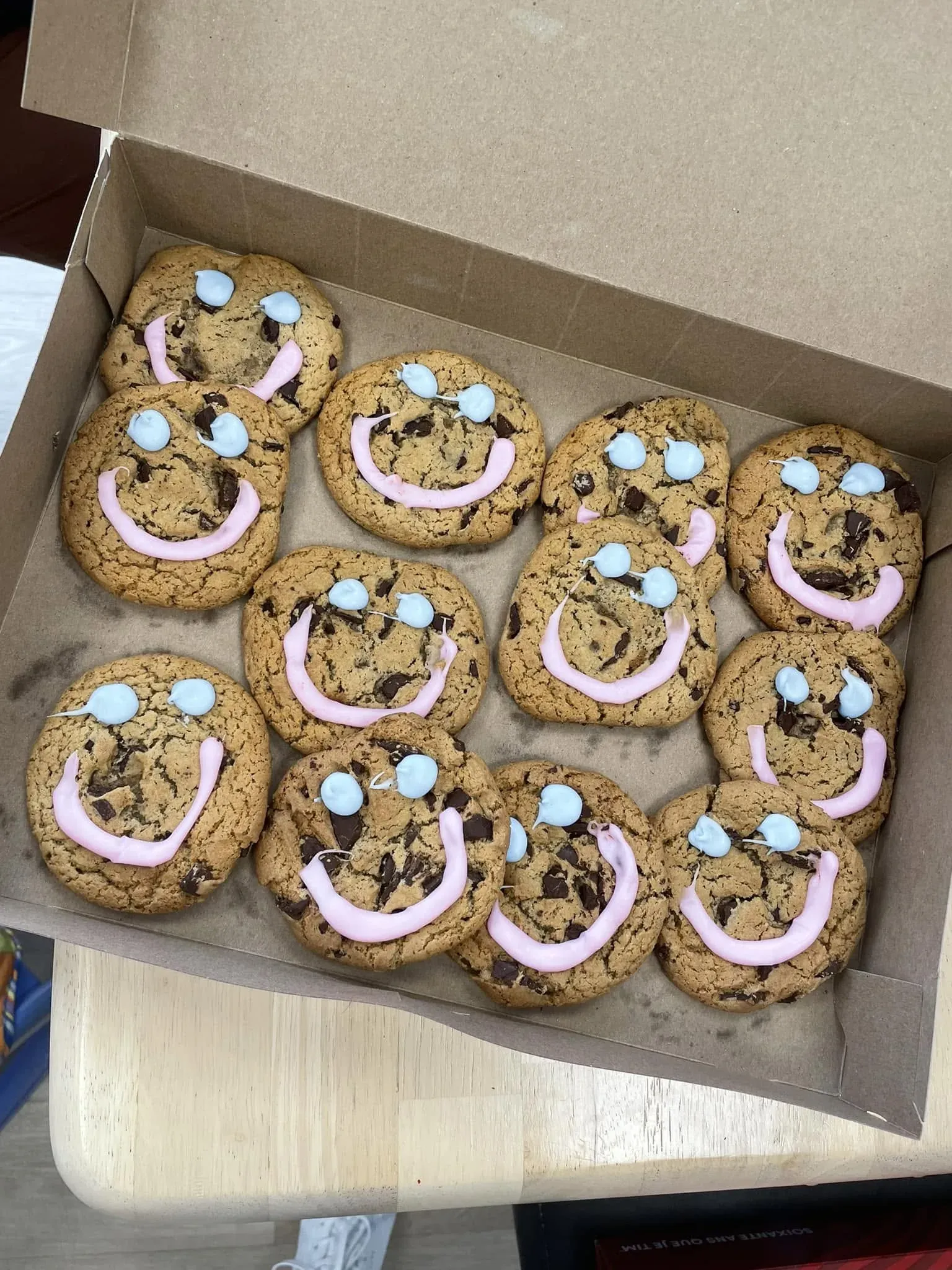 Une boîte de biscuits aux pépites de chocolat décorés en visages souriants, glaçage rose et yeux bleus.